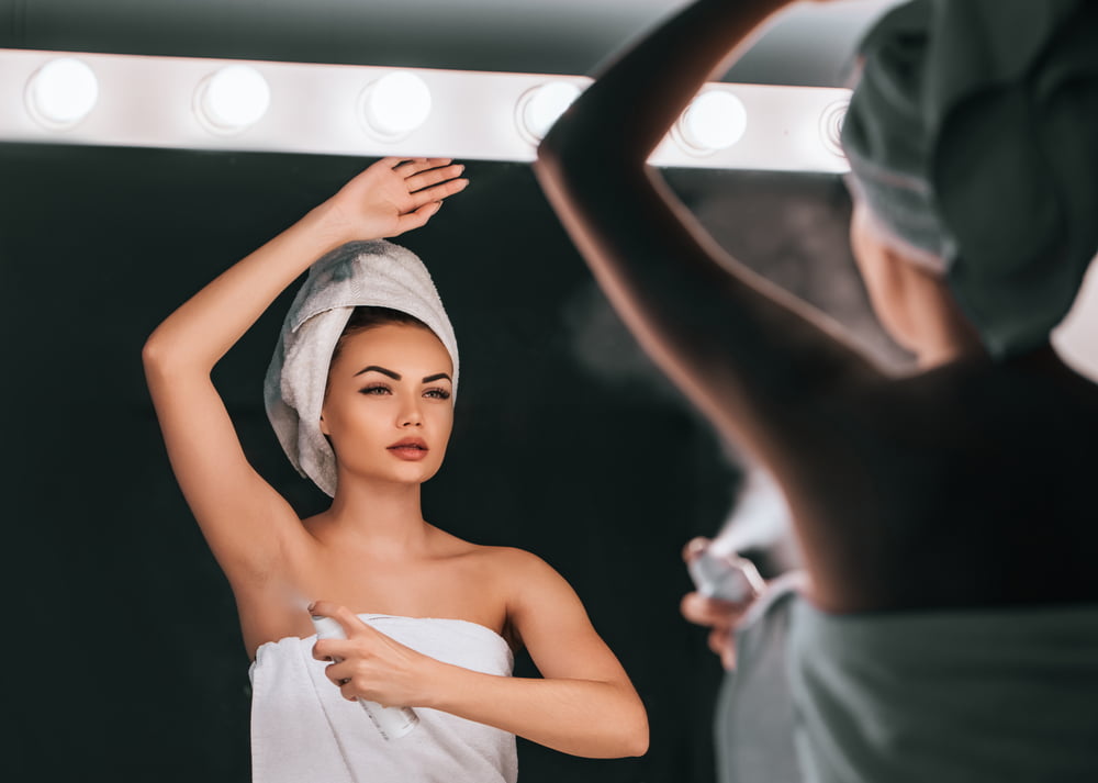 Attractive young woman in bathroom after shower is standing in front of mirror with deodorant in hands