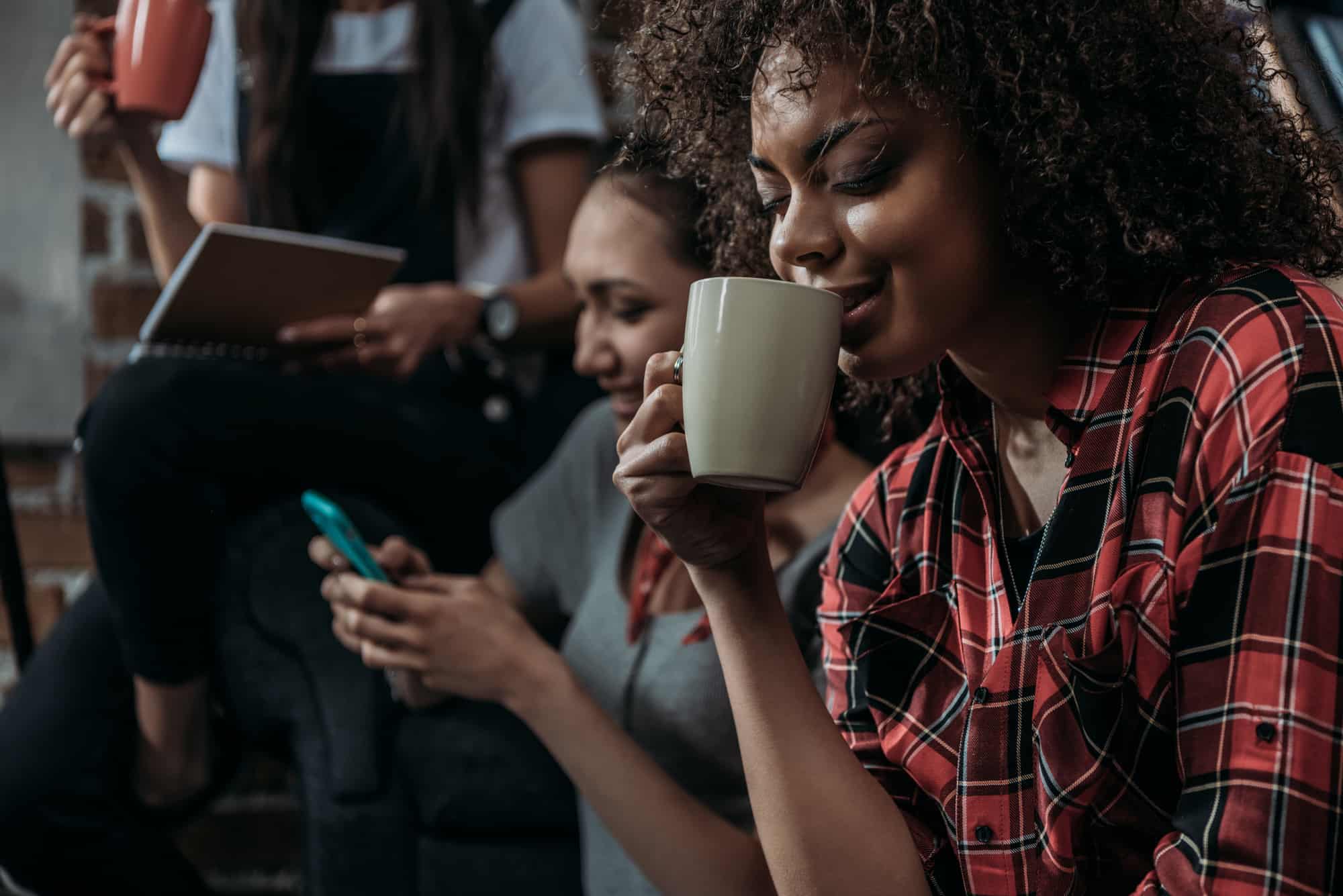 Happy young african american woman drinking coffee while friends using smartphone and holding cup behind