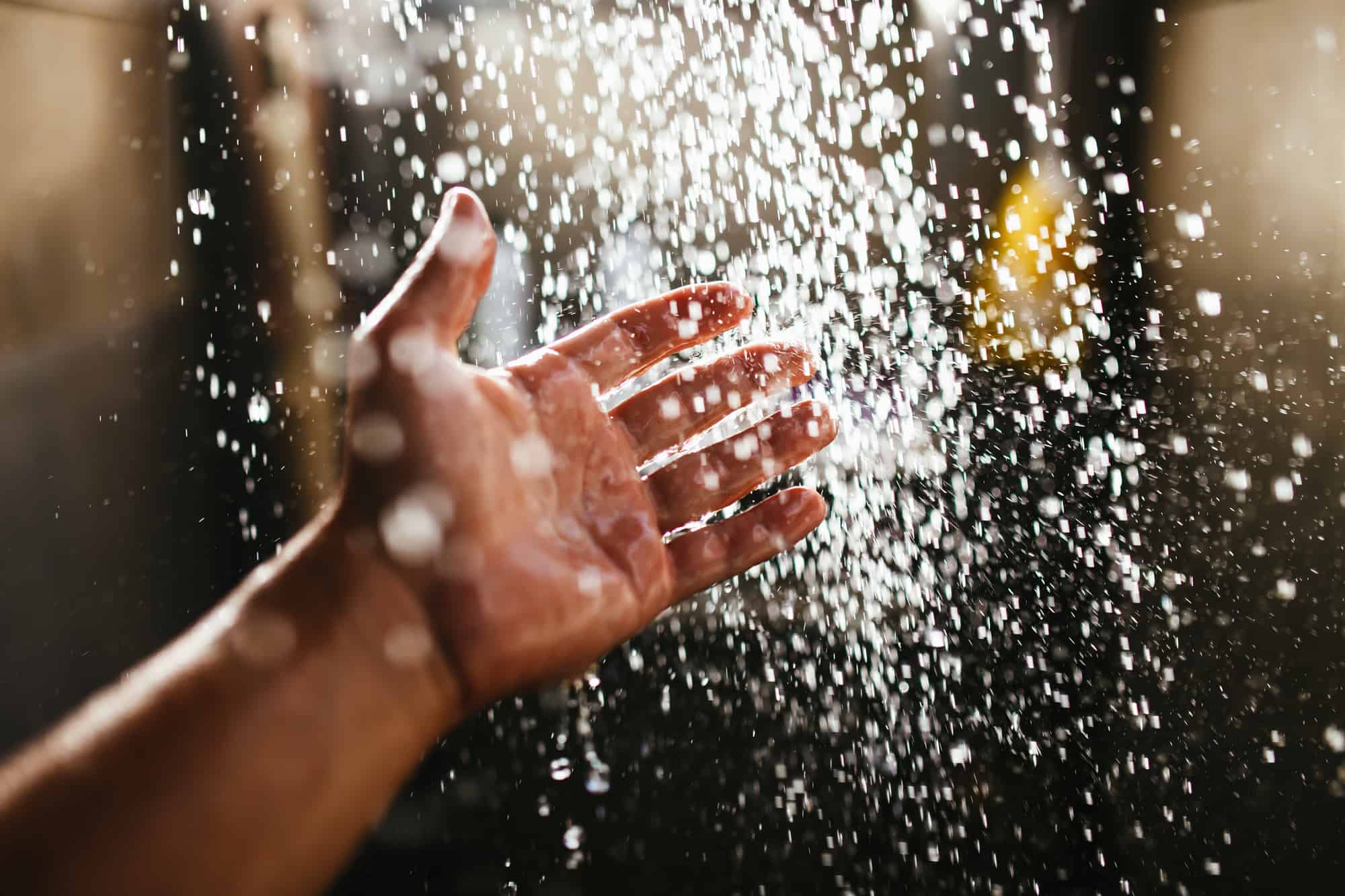 A man's hand in a spray of water in the sunlight against a dark background. Water as a symbol of purity and life