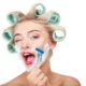 Cheerful woman has having cream over her face. She is shaving herself with razor. The girl is winking and laughing
