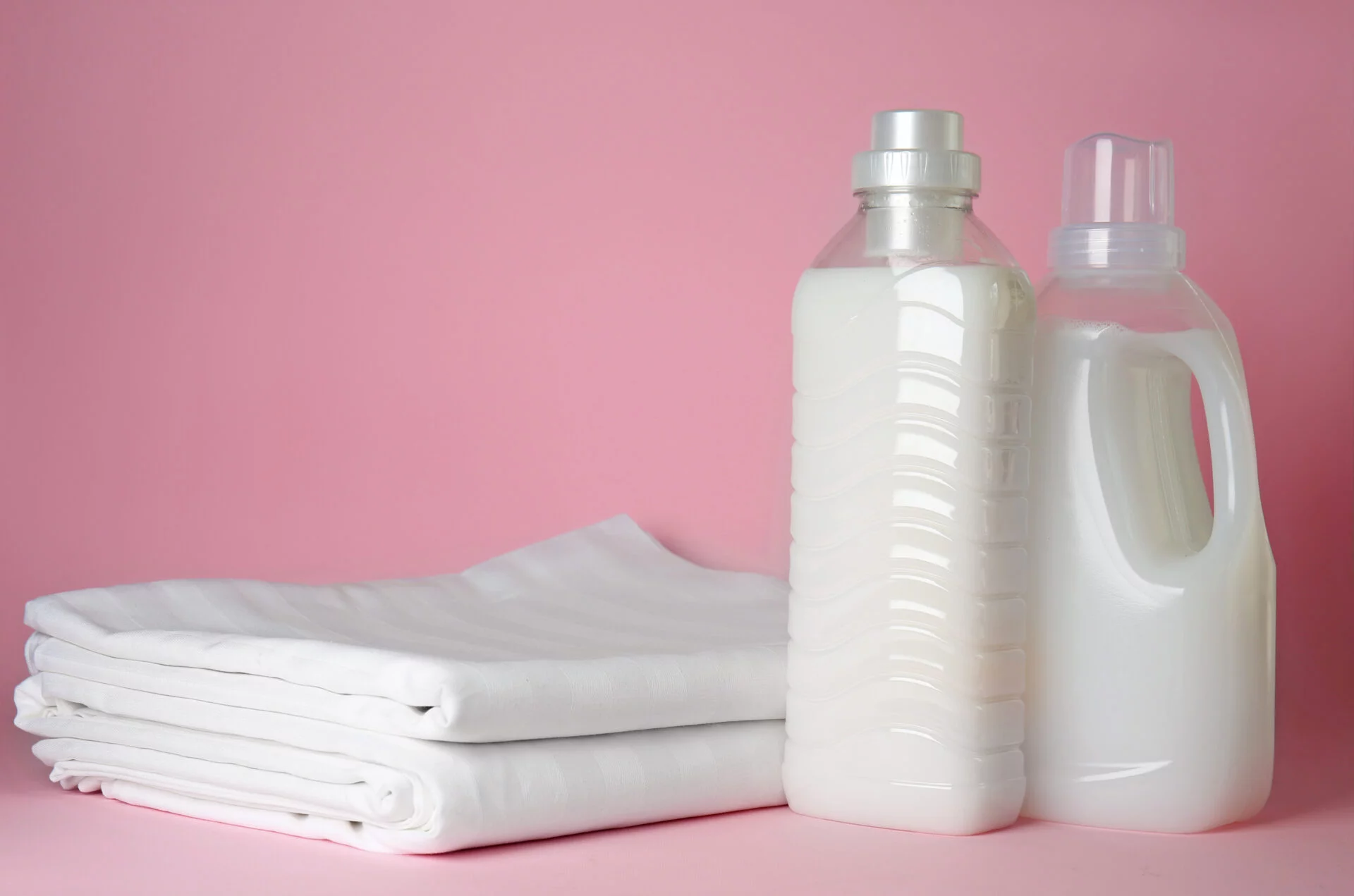 Stack of clean bed sheets and bottles with fabric softener on color background