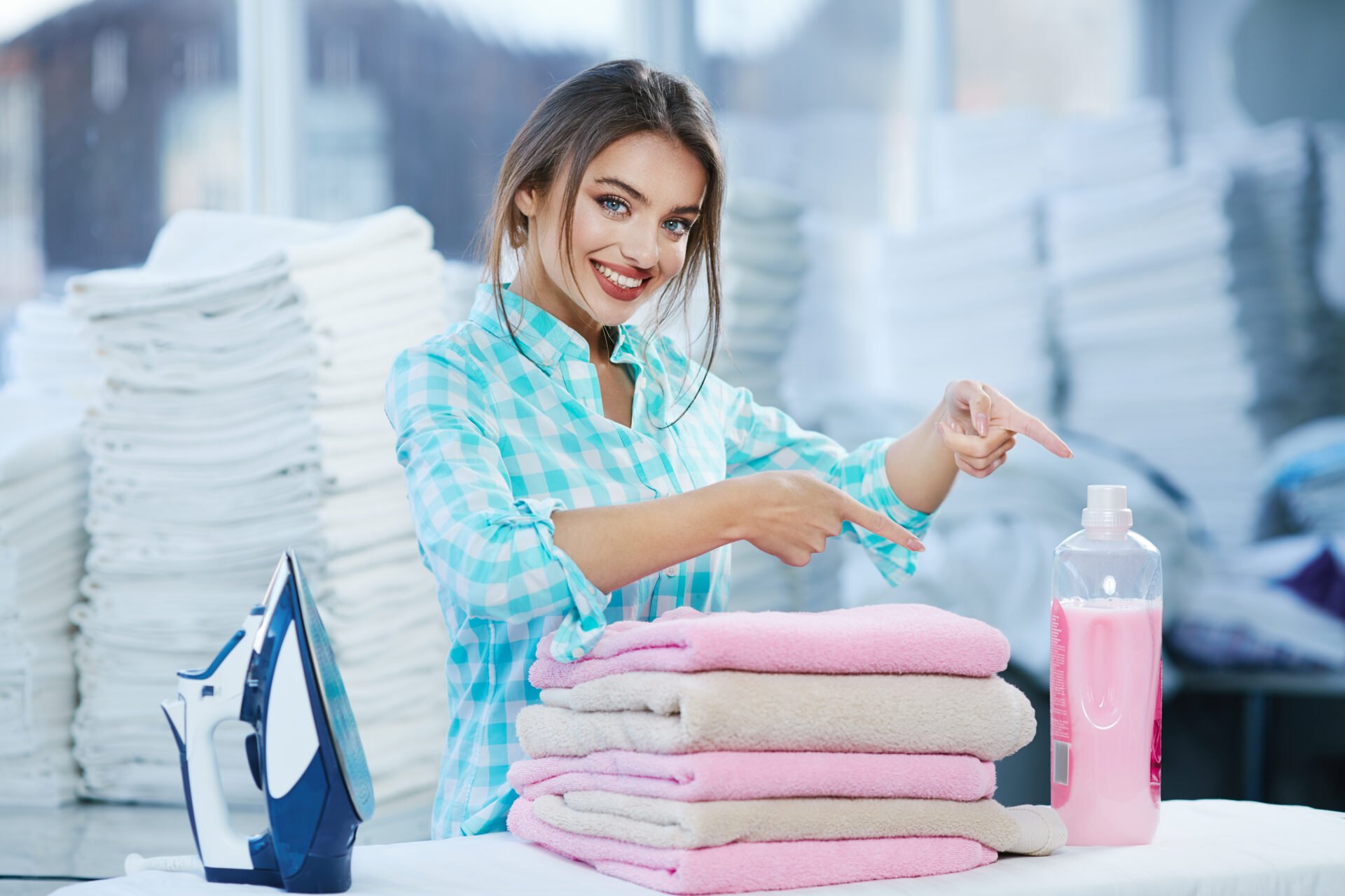 Woman standing near heap of rose towels and smiling. Woman near ironing board and detergent, heaps of white linen behind. Girl pointing at detergent