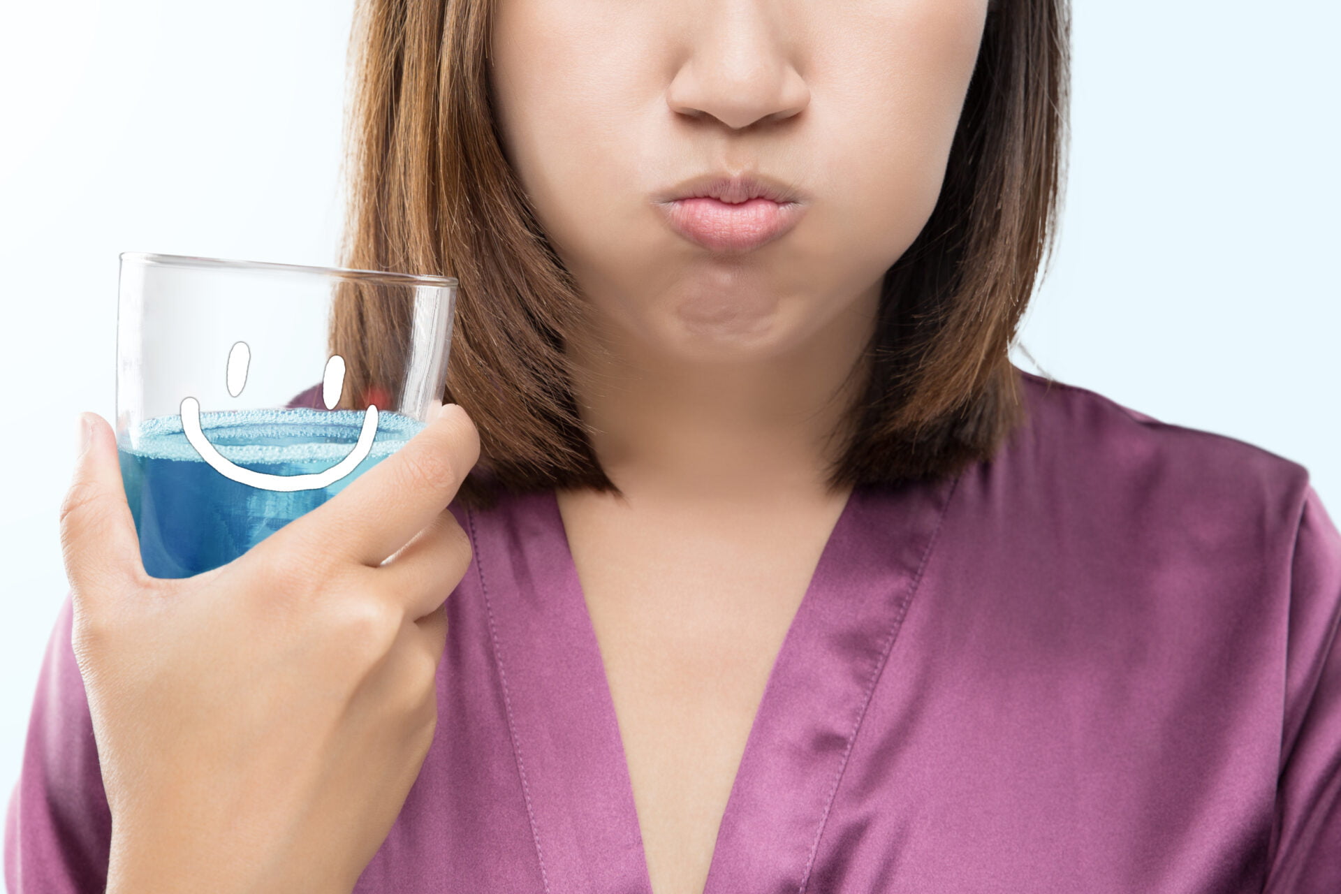 Asian woman rinsing and gargling while using mouthwash from a glass against gray background
