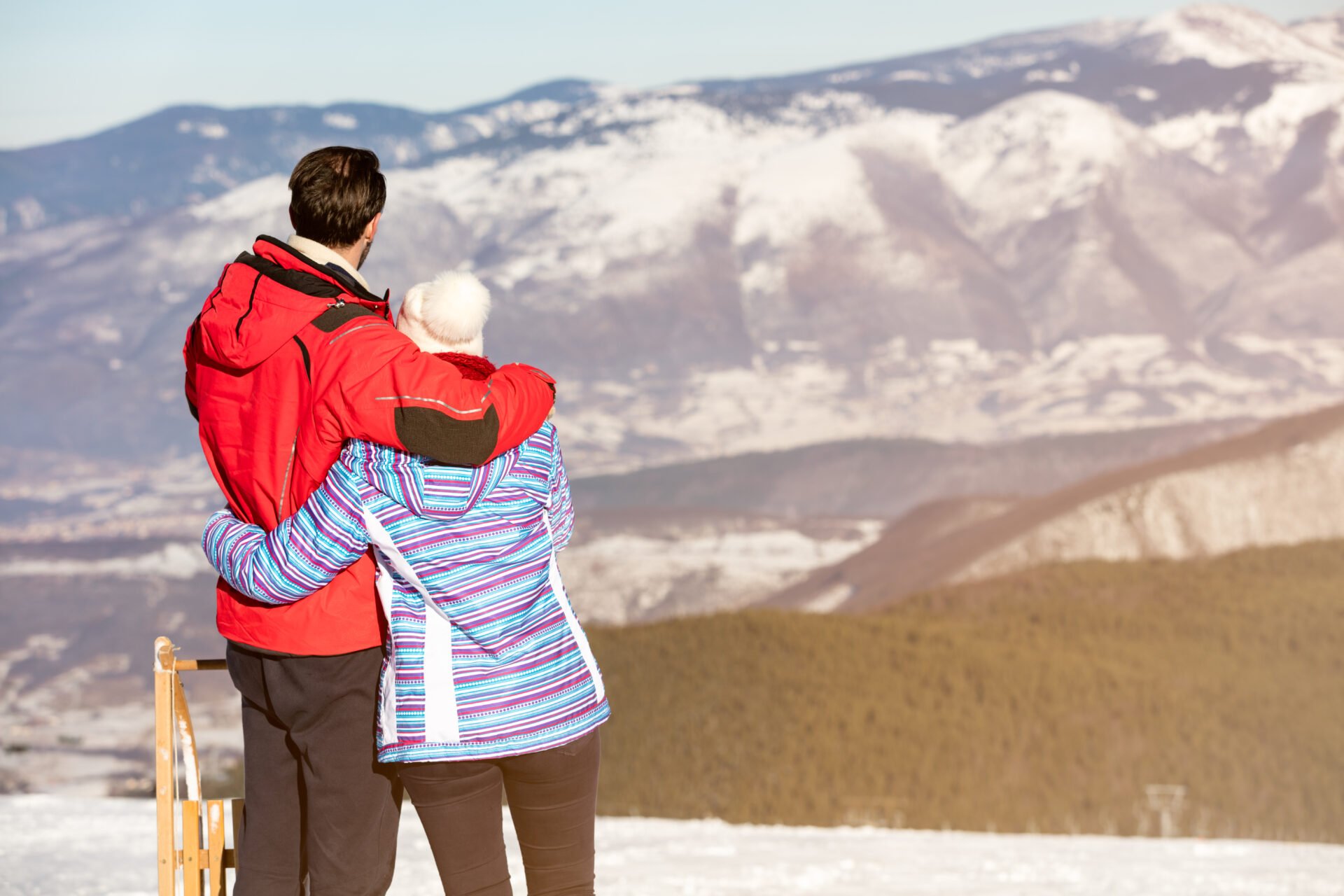 Rear view of a loving couple in fur hood jackets looking at snowed mountain range