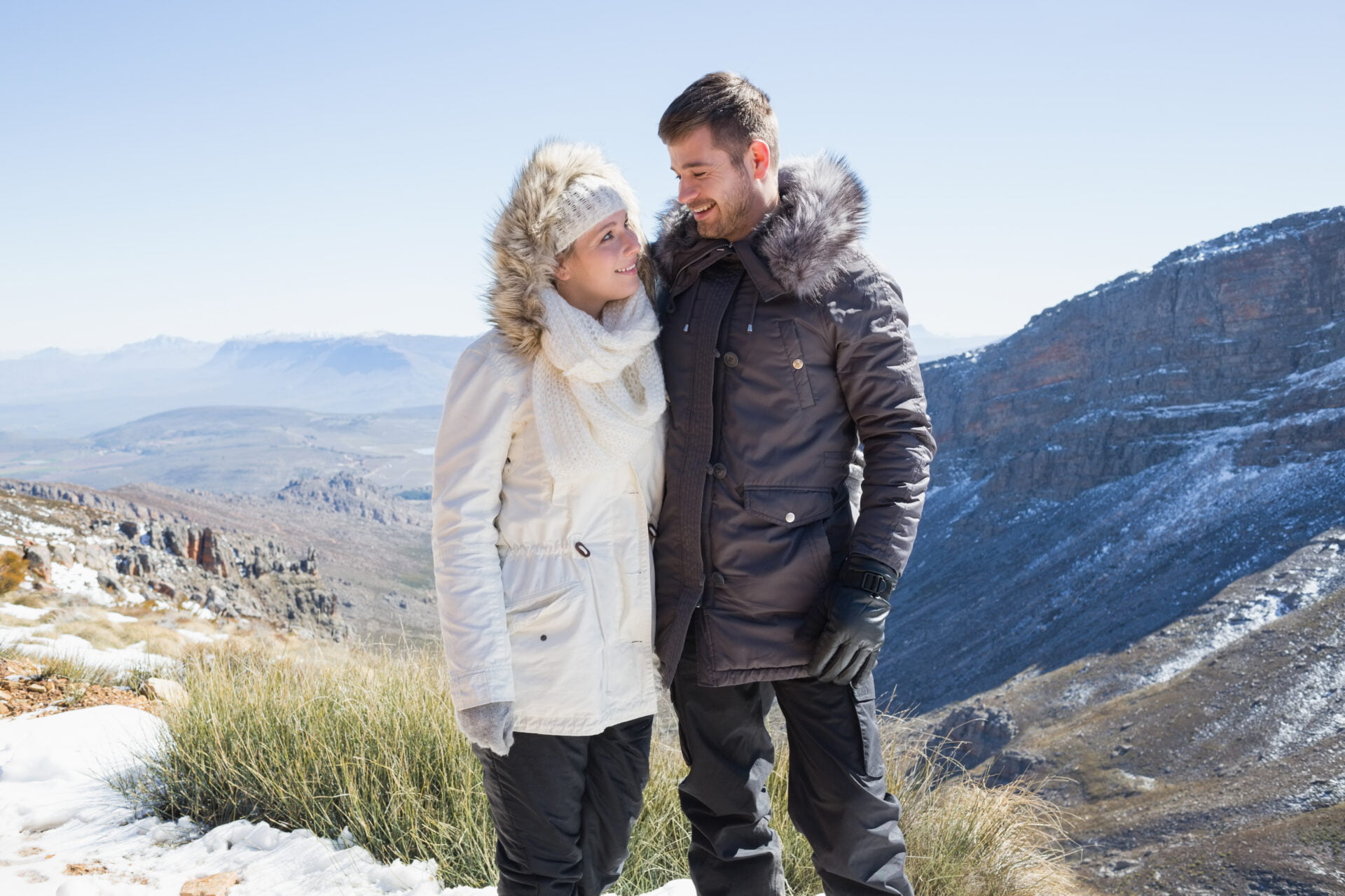 Smiling young couple in fur hood jackets against mountain range wearing PFAS free jackets