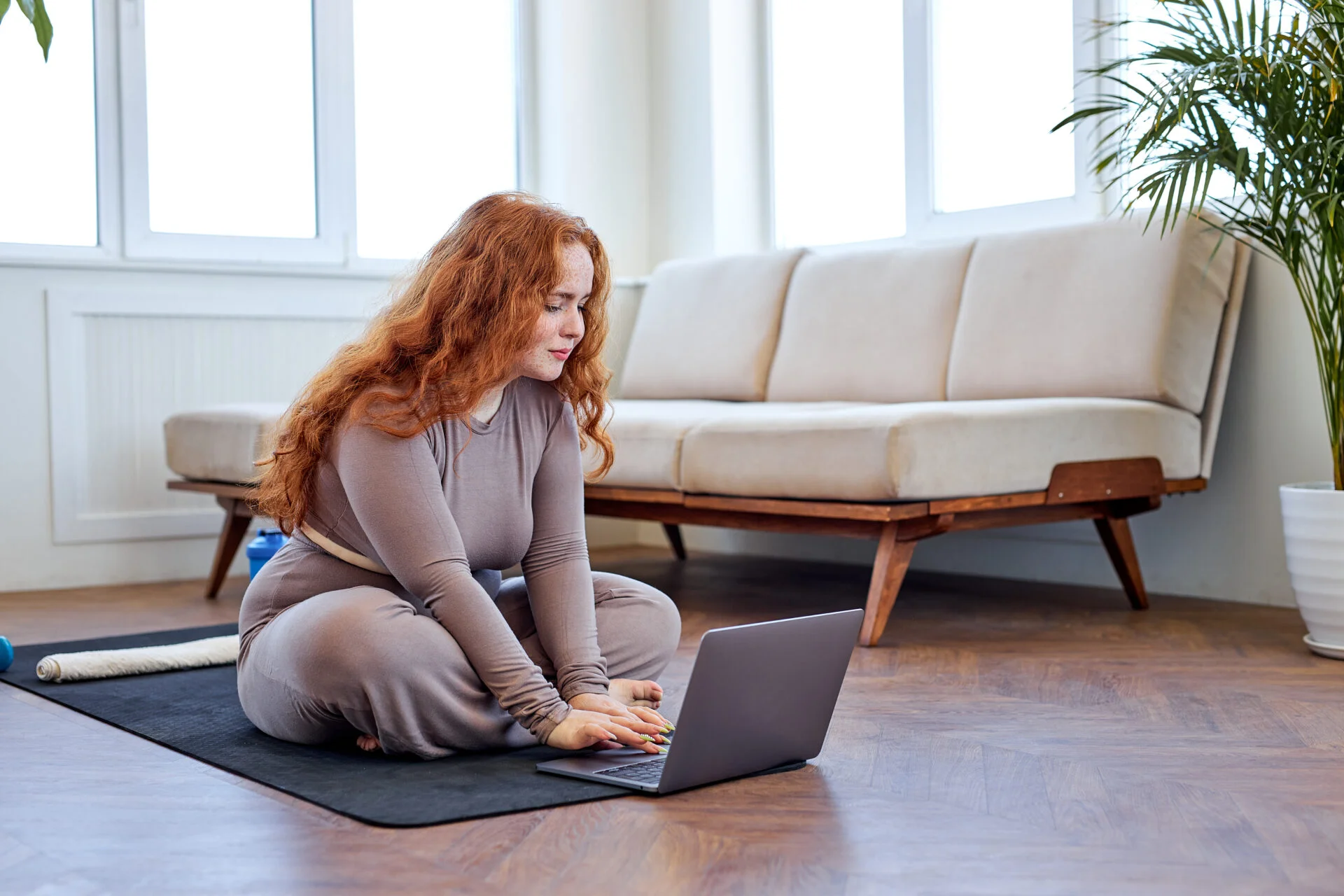 redheaded woman sitting on fitness mat at home in living room, using laptop, in sportswear