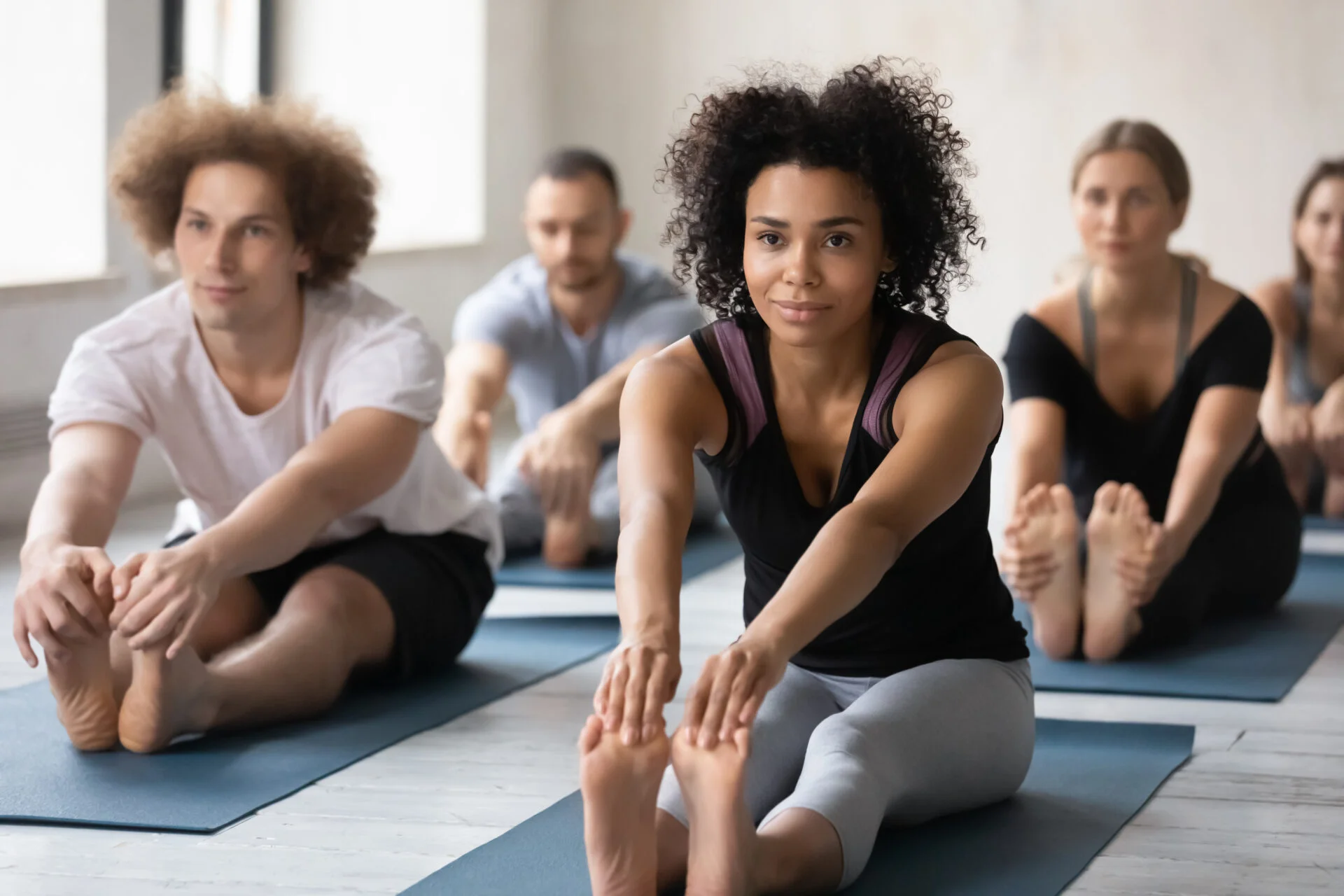 Wearing comfy stylish grey black color activewear group of people working out performing Seated Forward Bend exercise. Mixed-race woman and associates gather together for yoga training at sport club