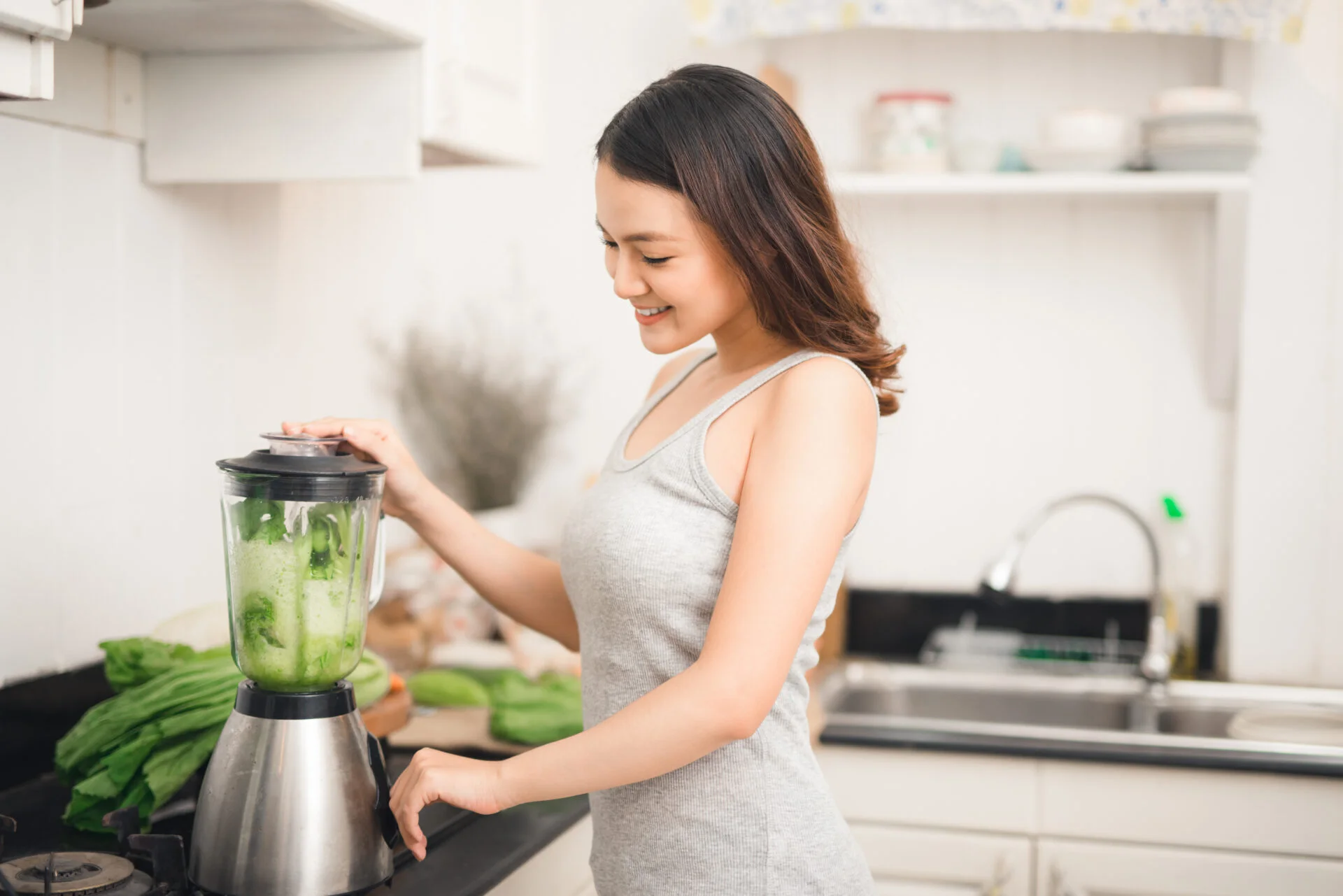 Smiling asian woman making smoothie with fresh vegetables in the blender in kitchen at home