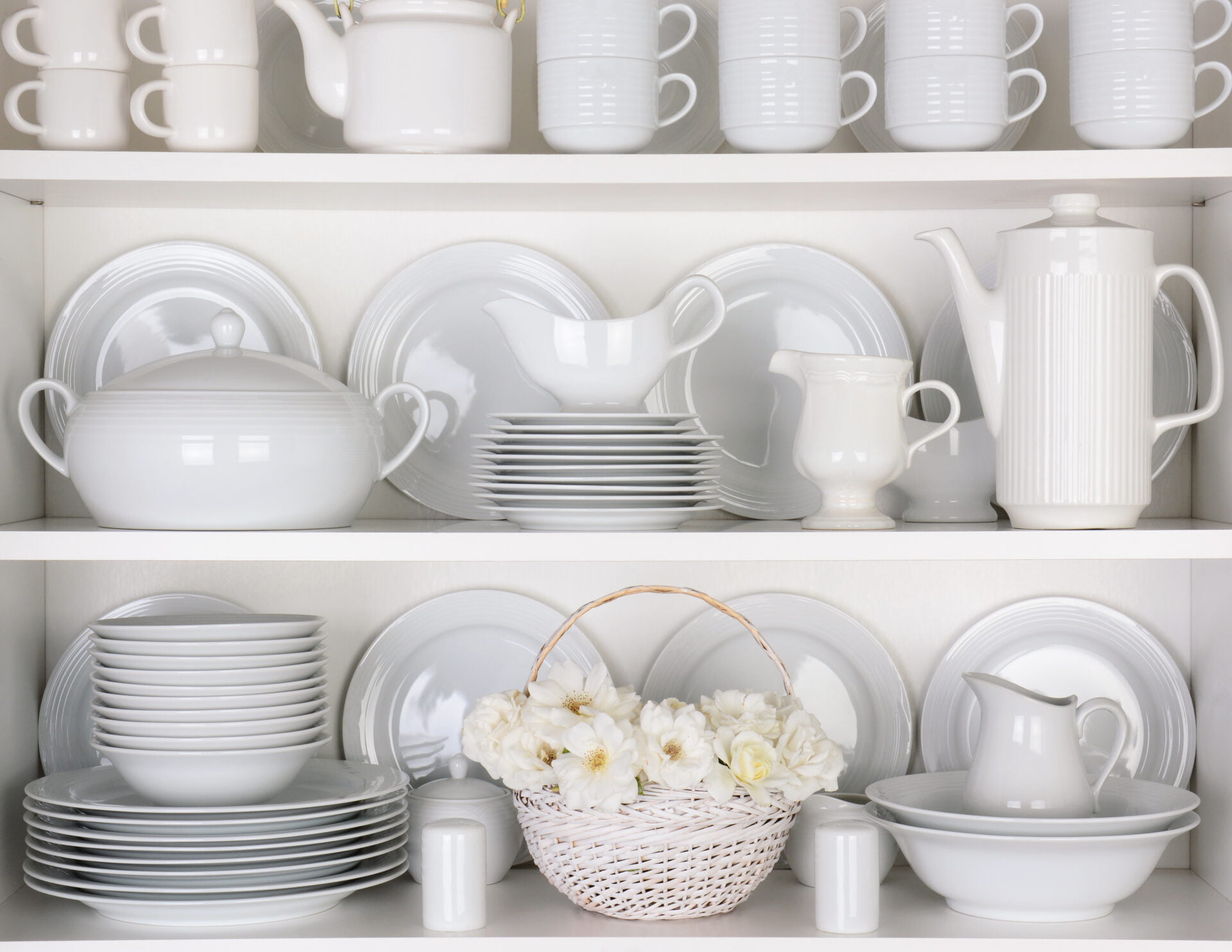 Closeup of white plates and dinnerware in a cupboard. A basket of white roses is centered on the bottom shelf. Items include, plates, coffee cups, saucers, soup tureen, tea pot, and gray boats