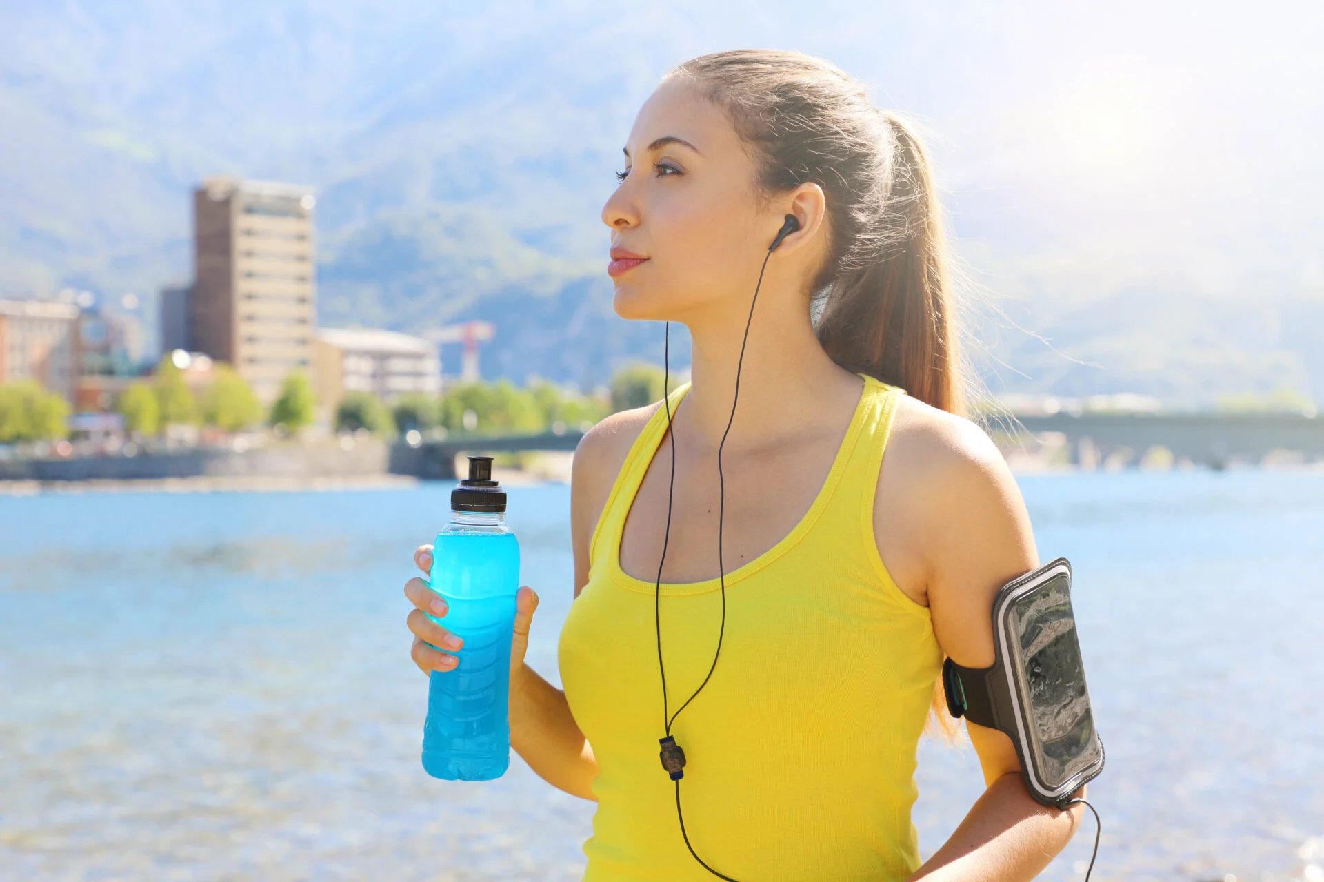Thirsty female athlete with armband for smart phone holding power drink and looking away outdoors.