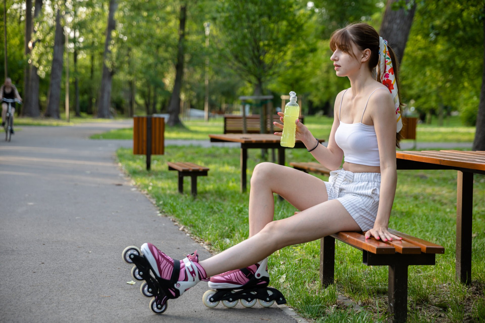 A young girl in shorts and a sports top sits on a bench. She is holding a bottle with a yellow isotonic drink. A figure riding a bicycle is visible in the background. Trees and lawns in the blurred background.