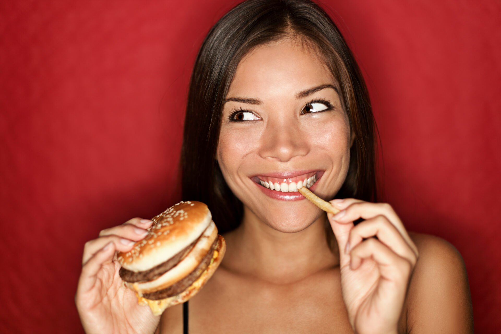 Woman eating burger and fries smiling. Beautiful mixed race asian caucasian female model on red background.