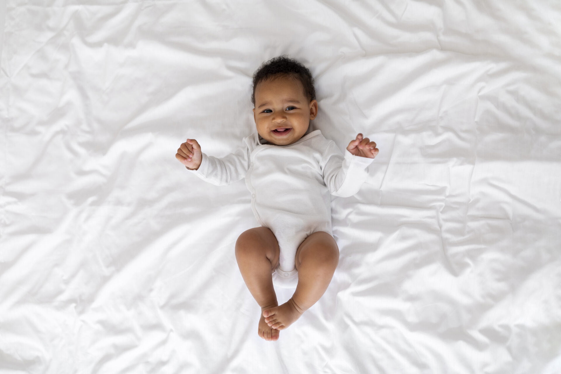 Portrait Of Cute Laughing Black Infant Baby Lying On Bed, Adorable Little African American Child In White Bodysuit Relaxing In Bedroom And Looking At Camera
