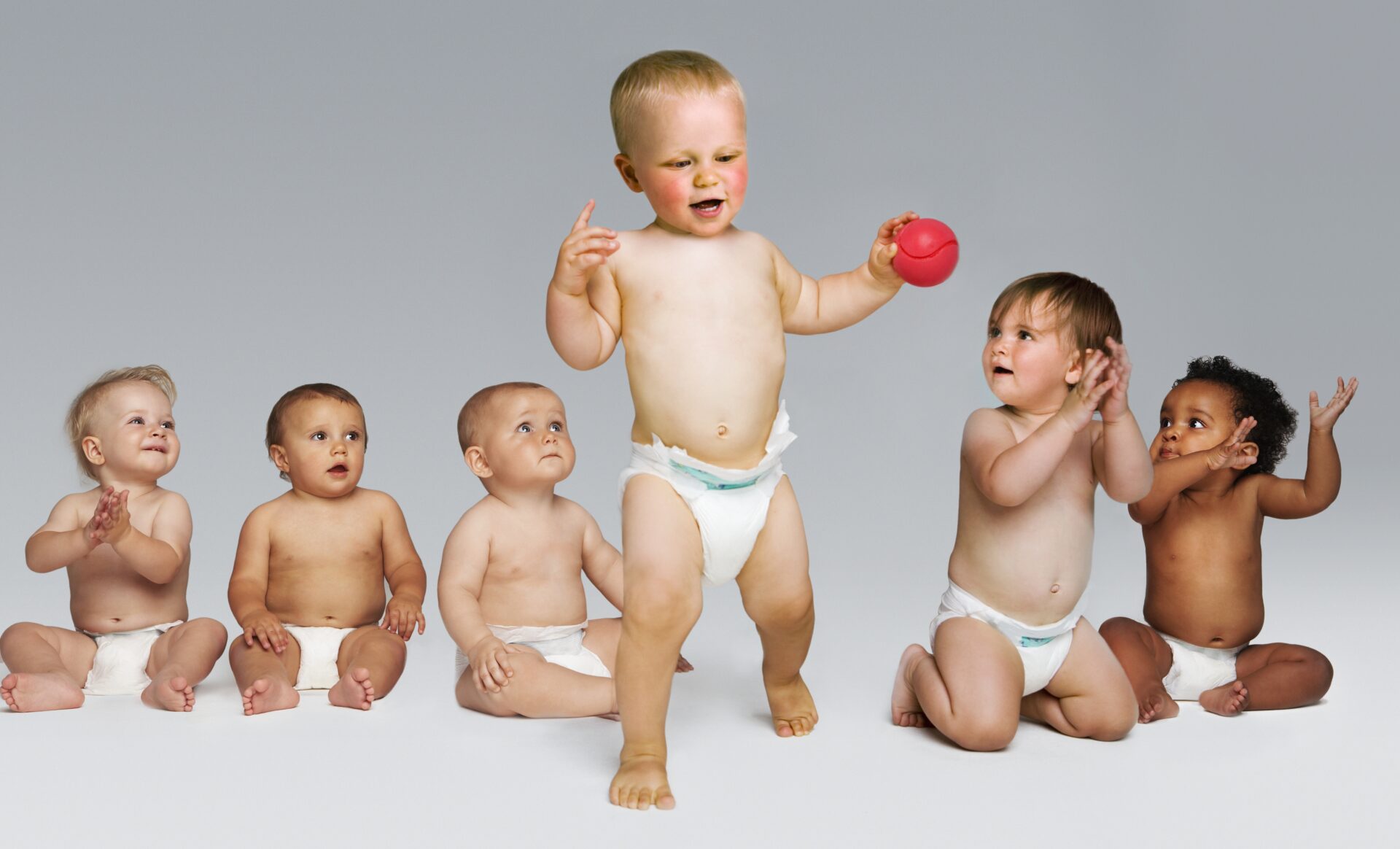 Babies sitting on ground in their diapers and one standing with a red ball
