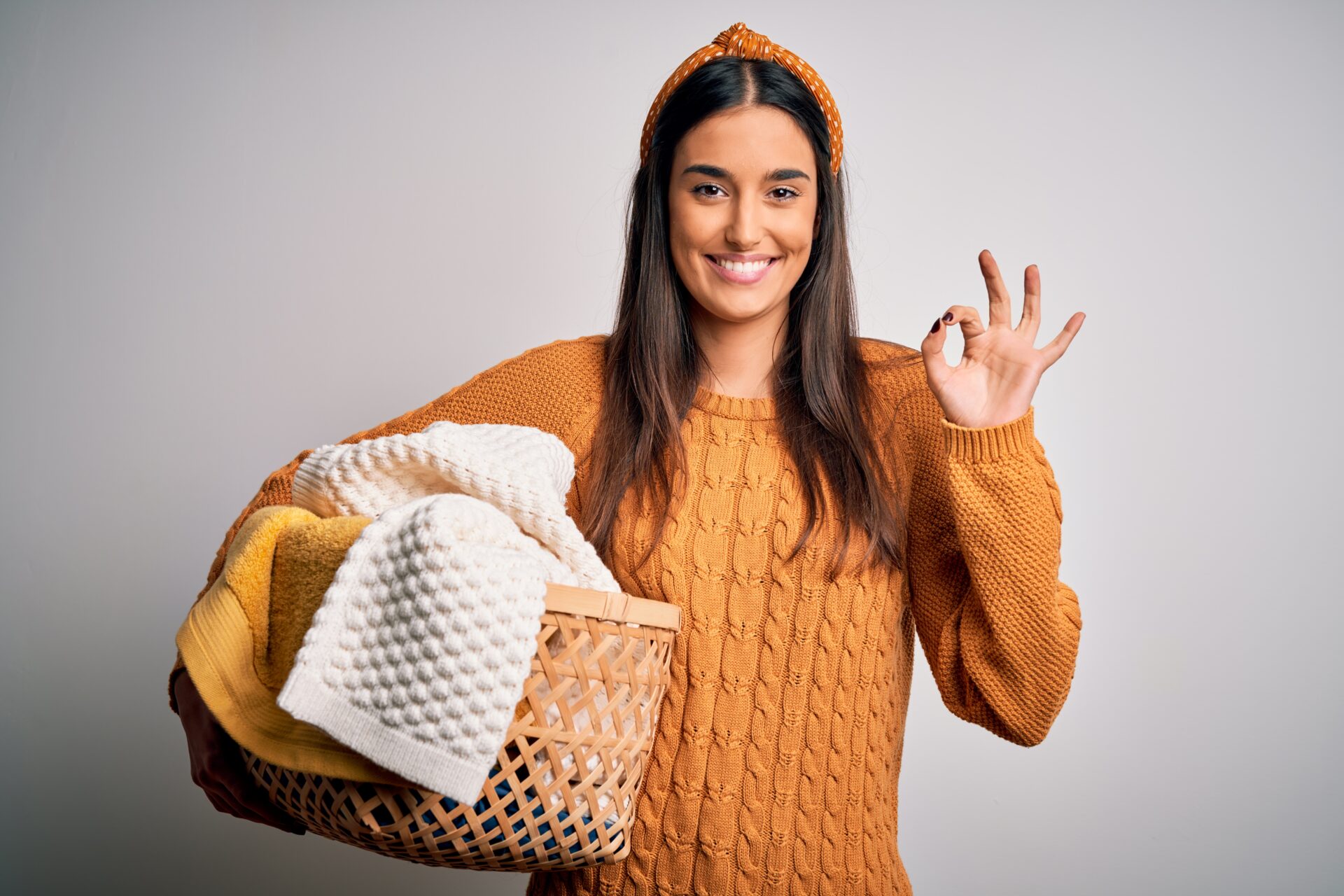 Young beautiful brunette woman doing housework chores holding wicker basket with clothes doing ok sign with fingers, excellent symbol