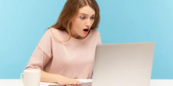Wow, unbelievable! Young woman with shocked expression looking at laptop screen with open mouth, talking on video call, online conference from home office. indoor studio shot isolated, blue background