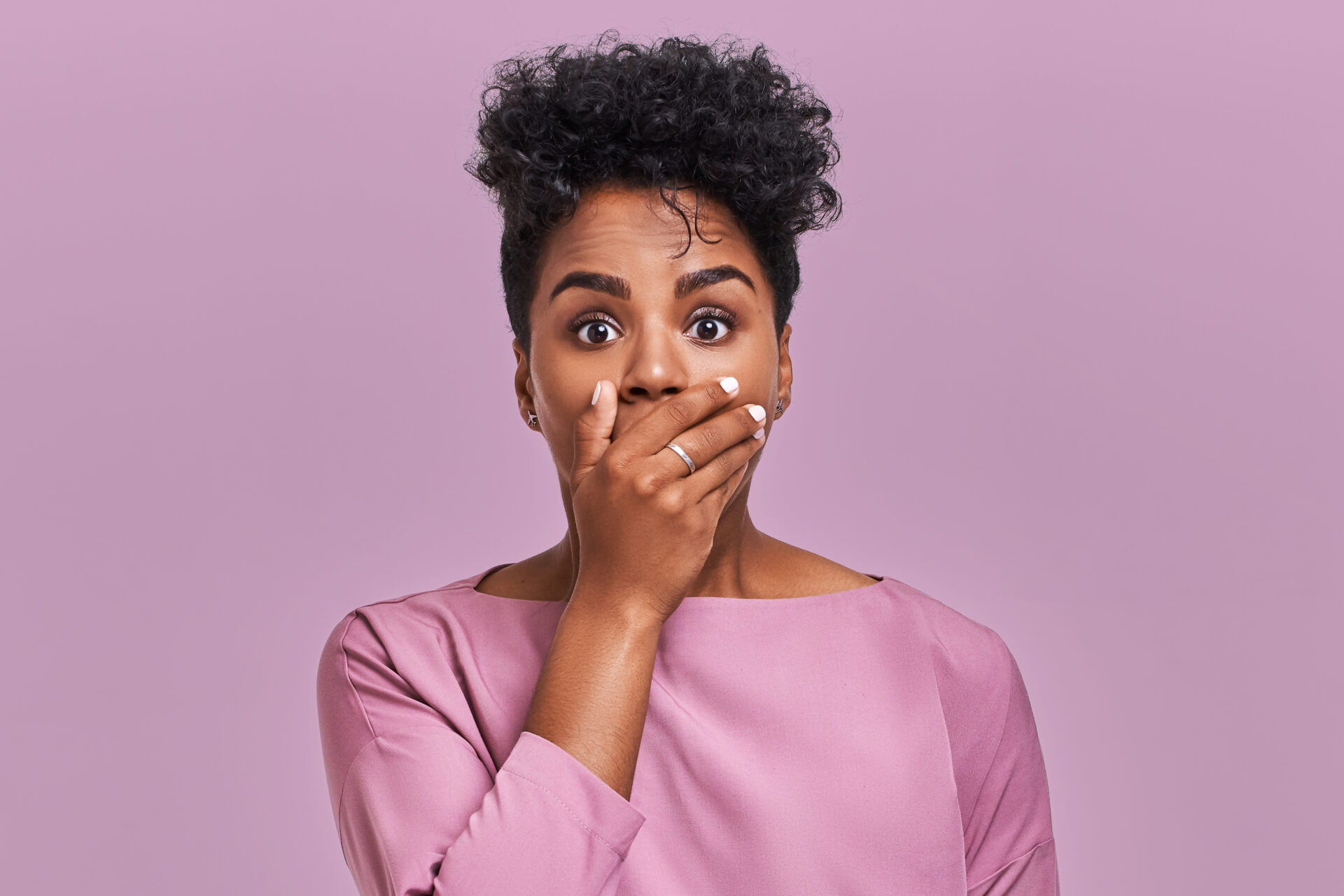 Horizontal shot of surprised African American woman covers mouth with both hands, tries to be speechless, looks with shocked expression, notices something strange, isolated over lavender blank wall.