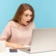 Wow, unbelievable! Young woman with shocked expression looking at laptop screen with open mouth, talking on video call, online conference from home office. indoor studio shot isolated, blue background